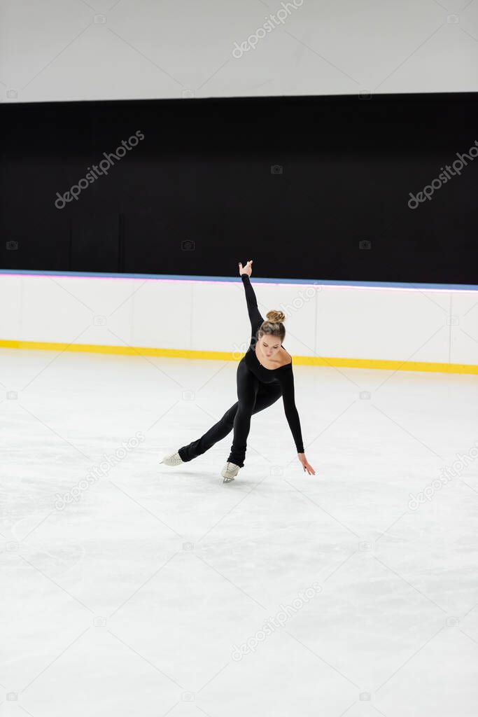 full length of young professional figure skater in black bodysuit skating with outstretched hand in ice arena 