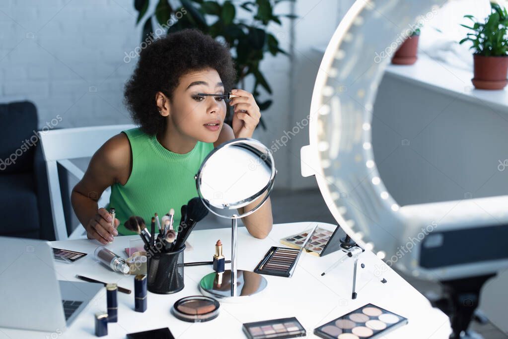 Pretty african american blogger applying mascara near mirror, gadgets and decorative cosmetics at home 