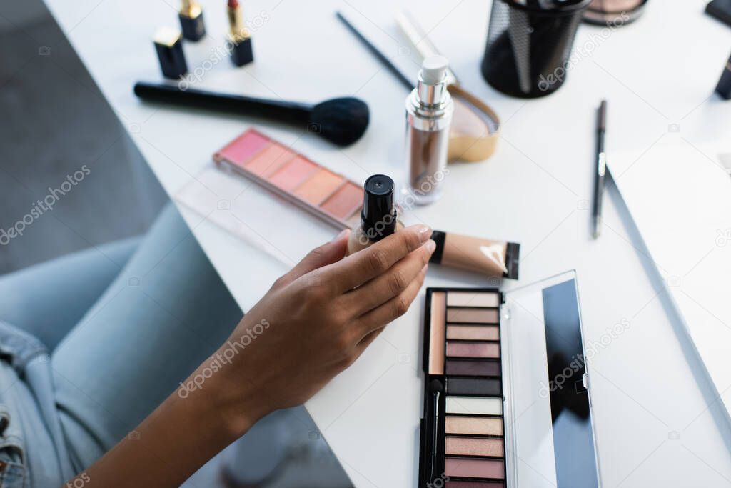 Cropped view of african american woman holding face foundation near blurred cosmetics on table 