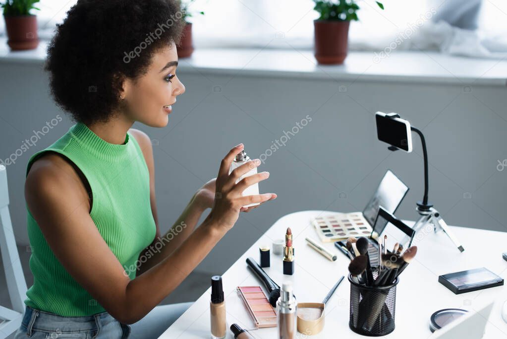 Side view of african american blogger showing perfume at smartphone near decorative cosmetics at home 