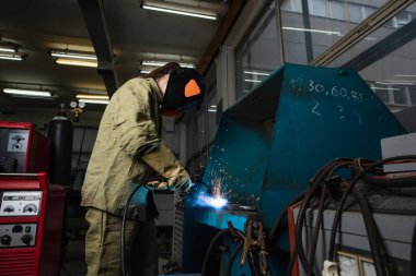 Welder in uniform and gloves working with welding torch near machine in factory  clipart