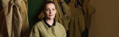 Young welder looking at camera near uniform in dressing room of factory, banner  clipart