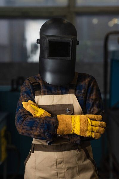 Welder in gloves, protective mask and overalls standing in factory 