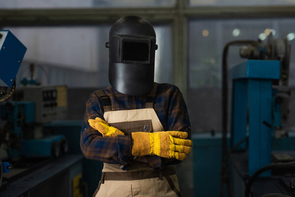 Welder in protective helmet with visor and gloves standing in factory 