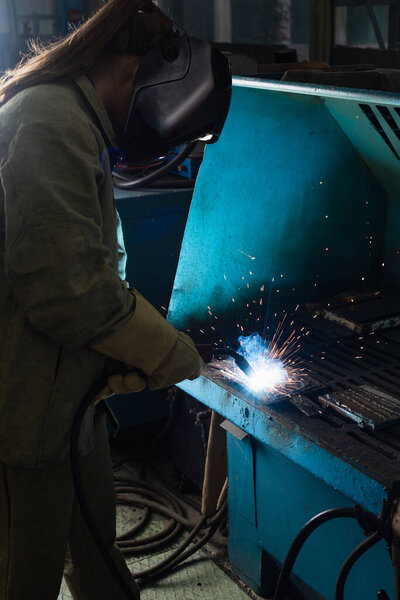 Young welder in uniform and protective mask working with welding torch in factory 