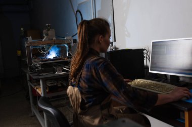 Fabrikada kaynak makinesinin yanında çalışırken bilgisayar kullanan esmer kaynakçının yan görüntüsü 