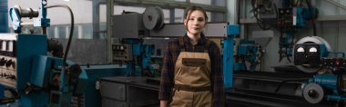 Brunette welder in overalls standing near welding machines in factory, banner  clipart