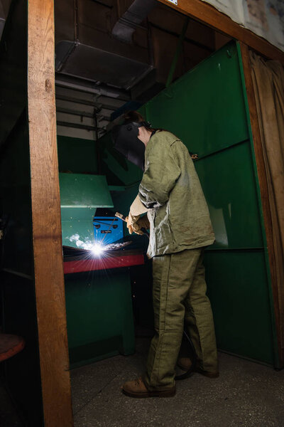 Full length of welder in protective uniform working with welding torch in factory 