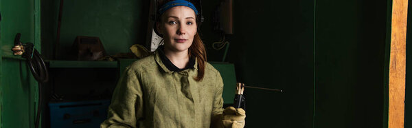 Brunette welder looking at camera while holding welding torch in factory, banner 