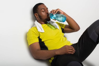 Yorgun Afrikalı Amerikalı sporcu spor şişesinden su içiyor ve gri renkte dinleniyor.