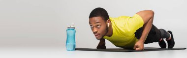 sportive african american man in earphones doing push ups on fitness mat near sports bottle on grey, banner