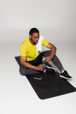Afrika kökenli Amerikan sporcusu spor şampanyasının yanında spor minderi ve gri renkte akıllı telefonuyla oturuyor.