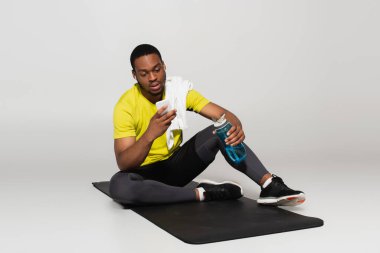 Tam boy Afro-Amerikan sporcusu spor minderinde oturuyor ve gri şişenin yanında akıllı telefon kullanıyor. 