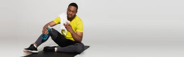 Tam boy Afro-Amerikan sporcusu spor minderinde oturuyor ve spor şişesinin yanında gri pankartta akıllı telefon kullanıyor.