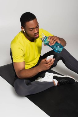 Afrikalı Amerikan sporcusu spor minderinde oturuyor ve gri şişenin yanında akıllı telefon kullanıyor. 