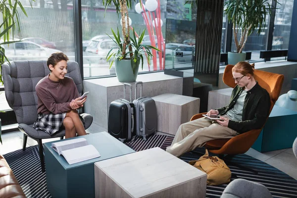 Genç çift otel lobisinde bavulların yanında akıllı telefon ve dijital tablet kullanıyor. 