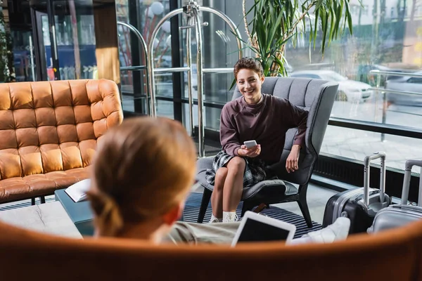 Smiling Woman Holding Smartphone Blurred Boyfriend Suitcases Hotel Lobby — стоковое фото