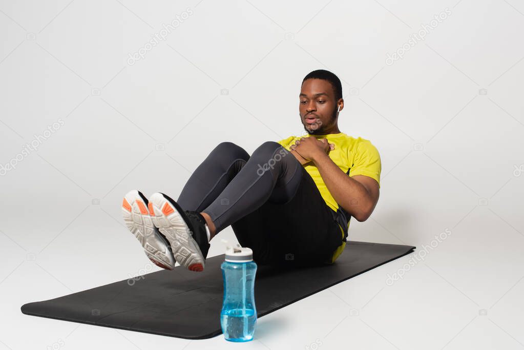 full length of sportive african american man in earphones doing abs exercise on fitness mat near sports bottle on grey