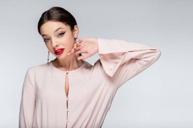 brunette woman in earrings and blouse looking at camera while posing isolated on grey clipart