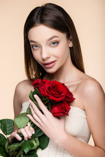 young woman with bare shoulders holding bouquet of red roses isolated on beige 
