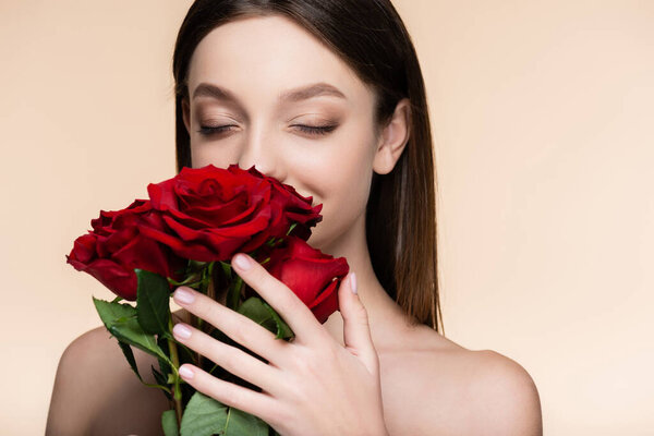 young woman with closed eyes smelling bouquet of red roses isolated on beige 