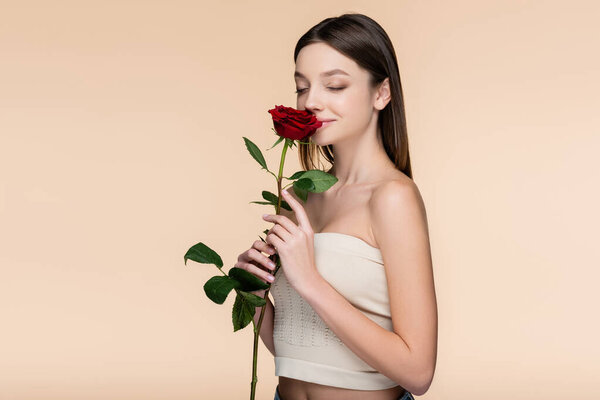 young woman with closed eyes smelling red rose isolated on beige 
