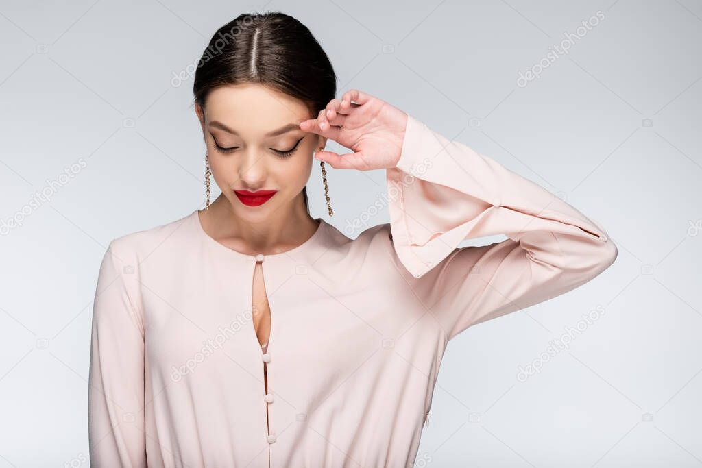 shy young woman in earrings and blouse looking down while smiling isolated on grey