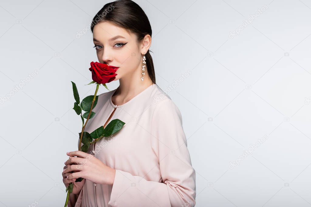brunette young woman in earrings and blouse smelling red rose isolated on grey