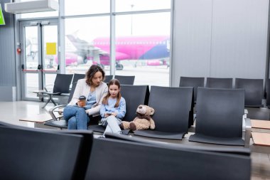 Havalimanı salonunda elinde kağıt bardakla annesinin yanında akıllı telefon tutan bir çocuk.
