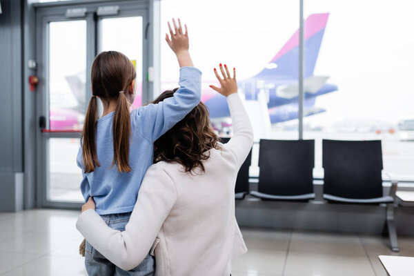 back view of mother and daughter waving hands while looking at plane in airport 