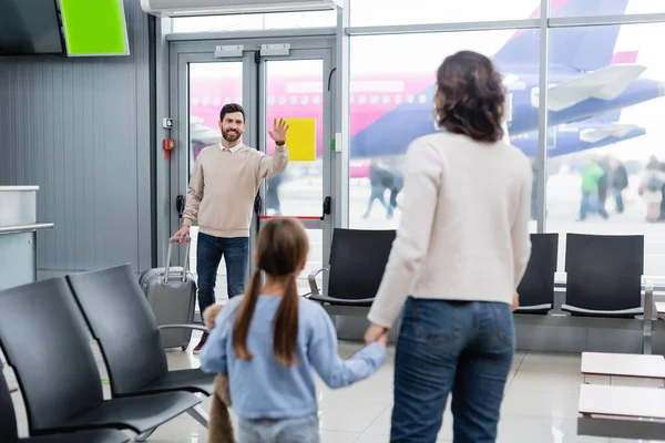 空港で妻と娘を見ながら手を振って荷物を持つ陽気な男 — ストック写真