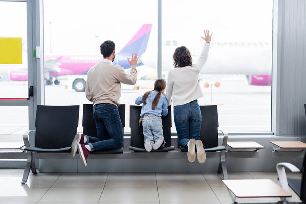 back view of family waving hands while looking at airplane through window in airport