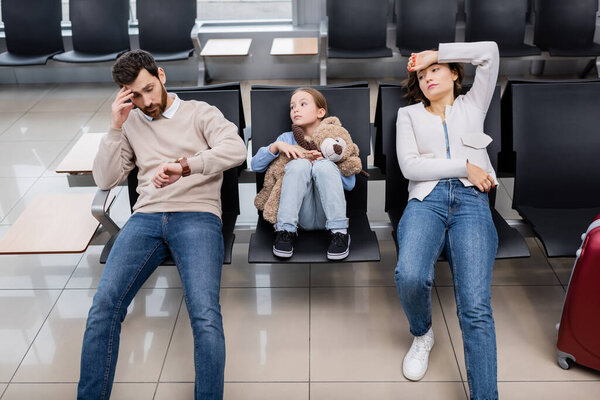 bearded man looking at watch near daughter and bored wife in airport 