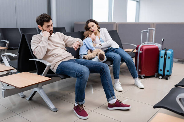 bearded man looking at watch near daughter and bored wife in airport lounge