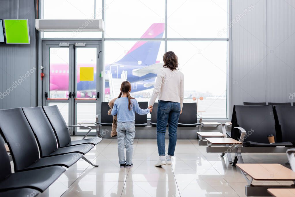 back view of mother and daughter holding hands while looking at plane in airport 