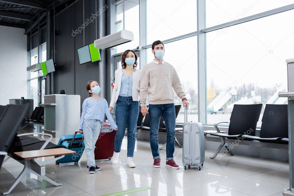 family in medical masks walking with luggage in airport lounge 