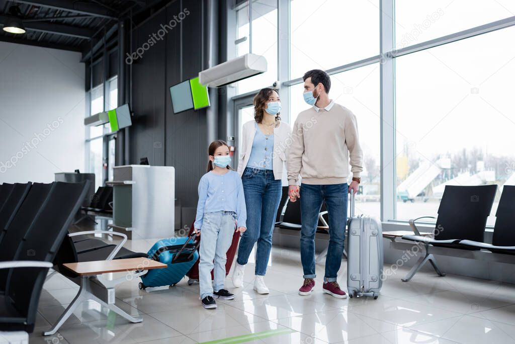 family in medical masks walking with baggage in airport lounge 