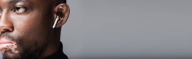 cropped view of blurred african american man with vitiligo listening music in earphone isolated on grey, banner