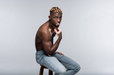 young african american man with vitiligo skin touching chin while looking at camera on high stool isolated on grey