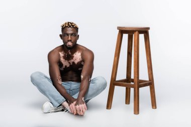 shirtless african american man with vitiligo skin sitting with crossed legs near wooden stool on grey