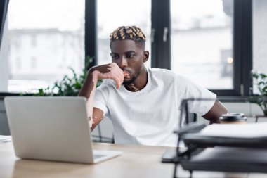 serious african american man with vitiligo thinking near blurred laptop in office