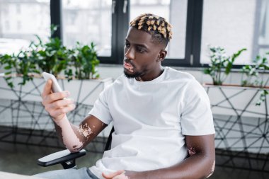 young african american man with vitiligo and trendy hairstyle looking at mobile phone while sitting in office