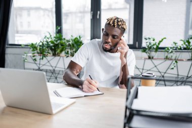 stylish african american man with vitiligo talking on smartphone and writing in notebook near blurred laptop