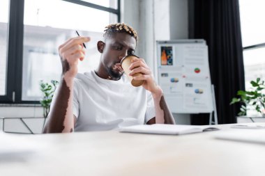 young african american man with vitiligo skin holding pen while drinking coffee near blurred notebook