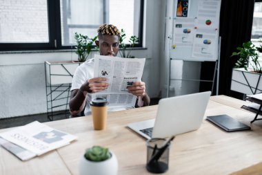 african american man with vitiligo reading newspaper near laptop and blurred flip chart in office