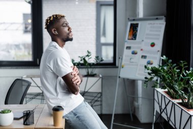 side view of african american man with vitiligo standing with crossed arms near coffee to go and blurred flip chart
