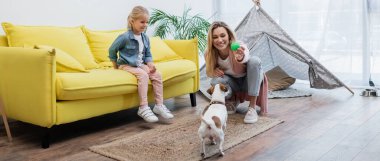Positive woman holding toy near jack russell terrier and daughter on couch at home, banner  clipart