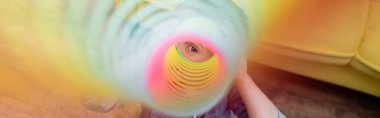 Child looking at camera through colorful slinky at home, banner 