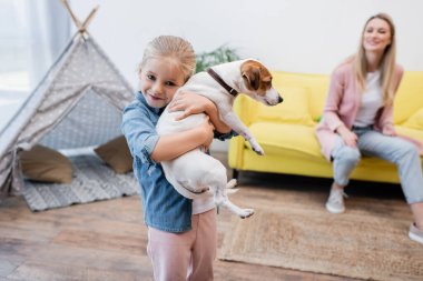 Happy kid hugging jack russell terrier near blurred mom at home 