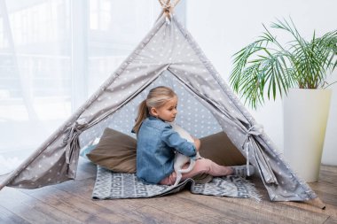 Girl hugging jack russell terrier in teepee near plant at home 